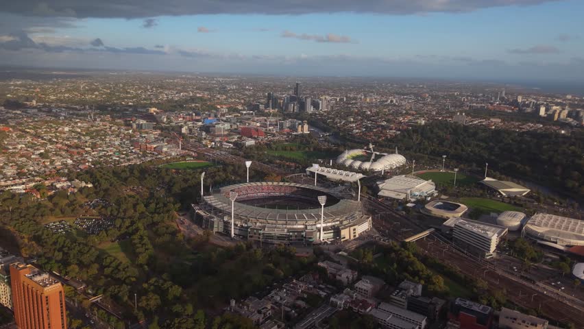 Sunset drone establishing orbit of Melbourne Cricket Ground with surrounding courts glowing in warm orange light