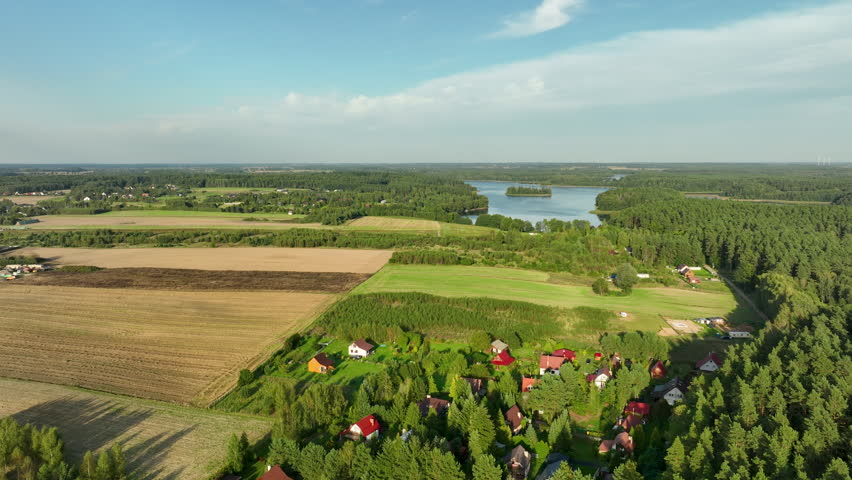 A sweeping aerial panorama of the Polish countryside, featuring patchwork fields of green and brown, a small cluster of colorful houses, and a forest-lined lake in the distance under a bright sky.