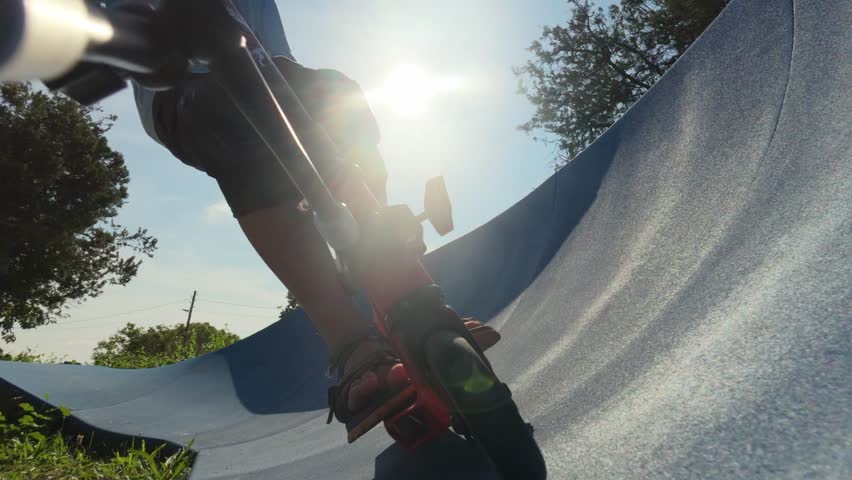 Close-up of a child riding a red scooter on a blue pump track, low angle view with summer trees and grass in the background