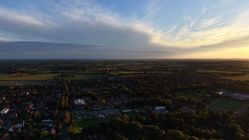 Cloudy golden fall evening in German village. Aerial wide shot. Colored trees and fields in late summer. Houses and Homes in peaceful town. Soccer field stadium of city. Sunset time.