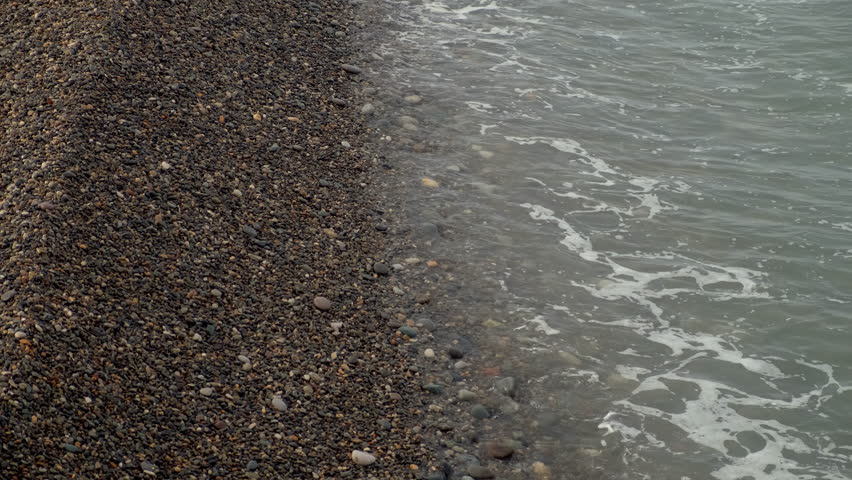Top view of long waves rolling onto pebble sea beach off-season time. Ocean waves softly washing over smooth pebbles, creating serene coastal landscape, peaceful atmosphere. Wild nature landscape.