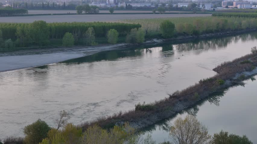 Sunset drone pan glides over the Po River with rowers slicing through golden water, riparian trees lining the floodplain, and Cremona’s skyline rising beyond cultivated terraces and alluvial banks