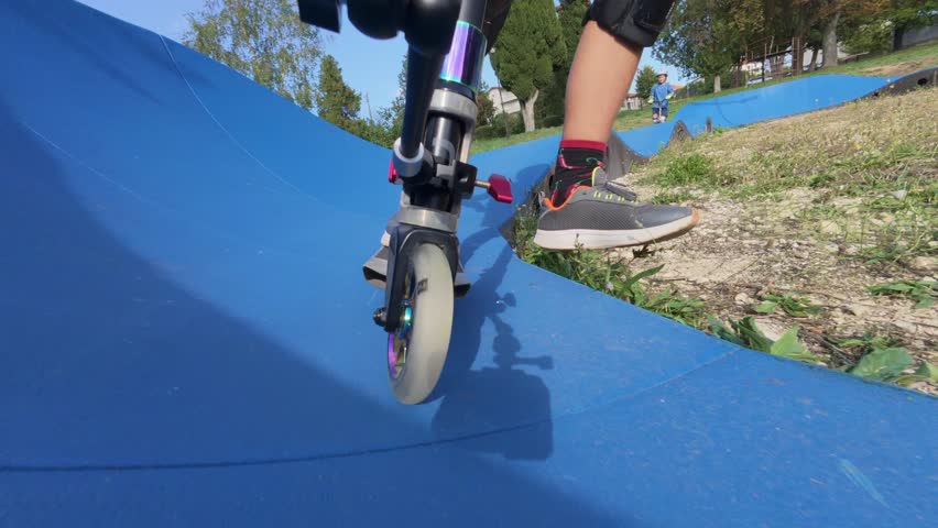 Close-up of a scooter wheel riding on a blue pump track in a park, with a child in protective gear enjoying outdoor extreme sport and active lifestyle