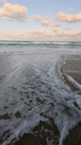 Slow motion sea waves washing sandy beach at sunset, foamy water reflecting clouds in the sky, peaceful seascape and natural ocean background