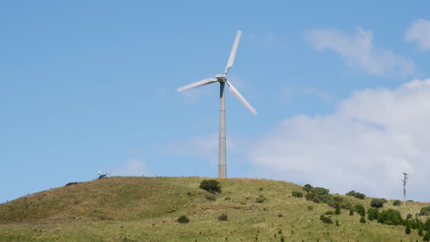 A single wind turbine generating power on top of a green hill with a clear sky in New Zealand