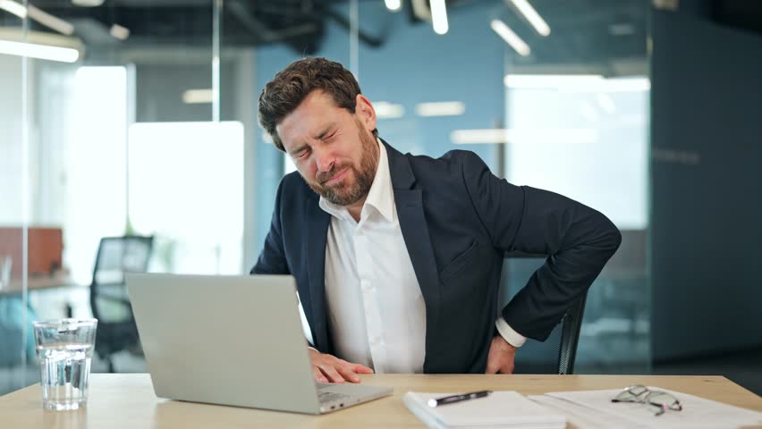 Handsome male freelancer, dressed in a suit, experiencing painful back discomfort in a modern office. His expression shows suffering from prolonged sitting.