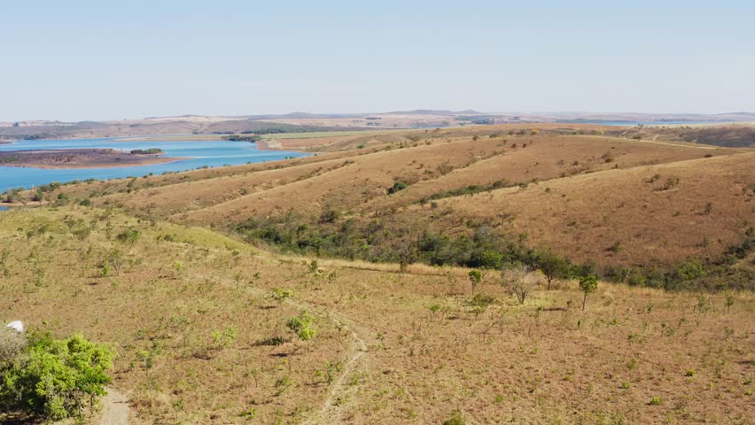 Drone orbits Grotas no Cerrado in Goiás revealing dry rolling hills, sparse vegetation, and a blue reservoir with islands showing biome zoning and terrain-water contrast in Brazil’s central savanna