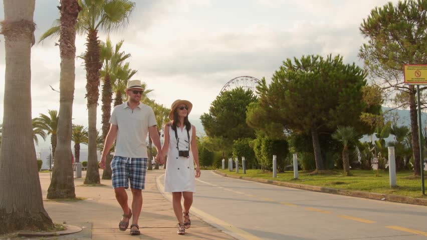 Young couple tourists walking hand in hand along Batumi promenade with palm trees and seaside view on sunrise, concept of tourism, vacation, lifestyle and romantic travel in Georgia