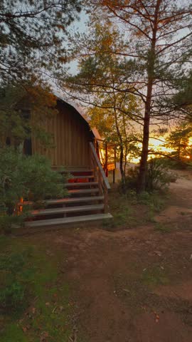 A warm and inviting vertical wide shot captures a rustic wooden cabin or tiny house nestled on the edge of a forest or hillside during the golden hour. The low evening sun casts long, dramatic shadows