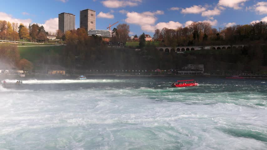 Boats for tourists float near the Rhine Falls with spray against the old town in Switzerland