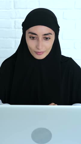 Vertical screen young muslim woman in black hijab smiling and waving during a video call on laptop from her modern home workspace
