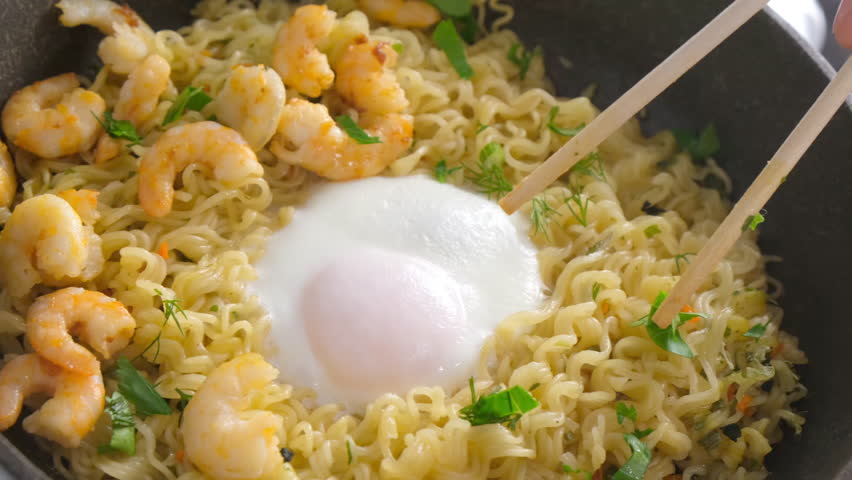 Chef breaking egg yolk with chopsticks near instant noodles, shrimps and herbs in frying pan. Preparing Asian dish. Cook fresh food domestic homemade recipe. Culinary, cuisine, fry noodles.