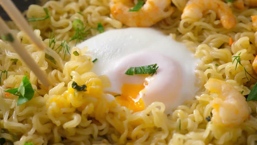 Chef breaking egg yolk with chopsticks, instant noodles, shrimps and herbs in frying pan. Preparing Asian dish. Cook fresh food domestic homemade recipe. Culinary, cuisine, fry noodles.