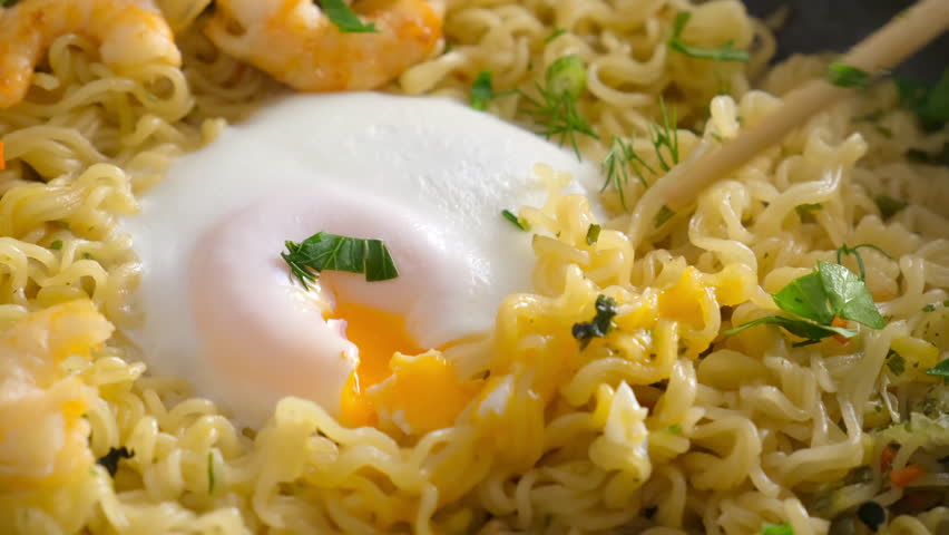 Chef breaking egg yolk with chopsticks near instant noodles, shrimps and herbs in frying pan. Preparing Asian meal dish. Cook fresh food domestic homemade recipe. Culinary, cuisine, fry noodles.