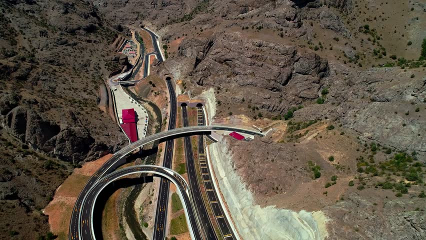 Aerial footage of a modern highway interchange with tunnels, bridges, and viaducts in a mountainous region. Advanced transportation infrastructure reflects engineering excellence.