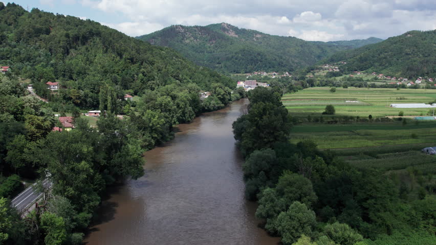Drone view of brown muddy river winding through farmland fields and forested hills