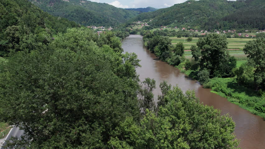 Aerial shot of muddy brown river flowing through valley with forest and farmland