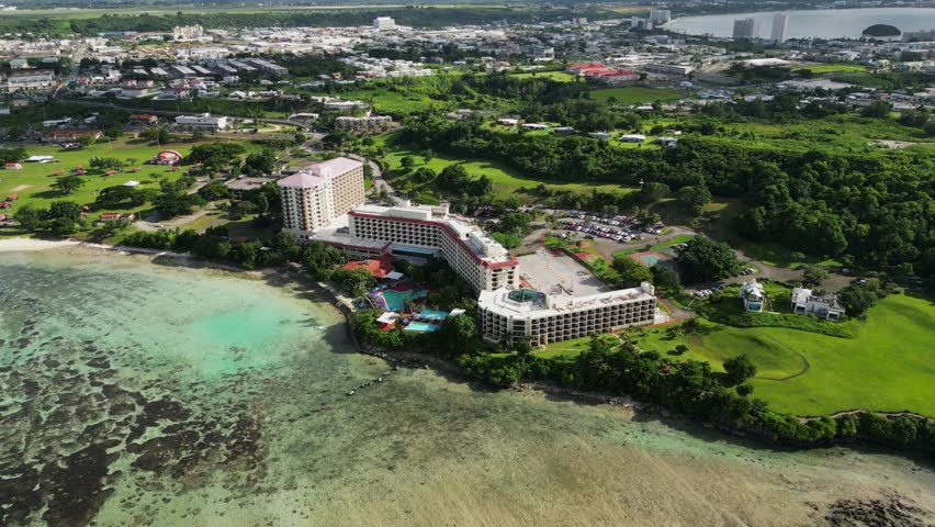 Picturesque view of coastal hotel resort with lush greenery facing shallow ocean reef at tropical island Guam, USA - aerial orbit