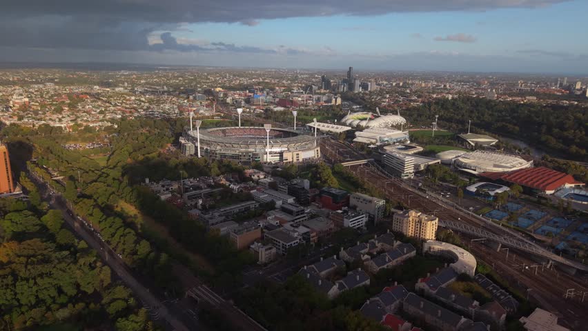 Aerial establishing at golden hour of Melbourne Cricket Ground and Tennis Courts with glowing sky backdrop, pullback panoramic