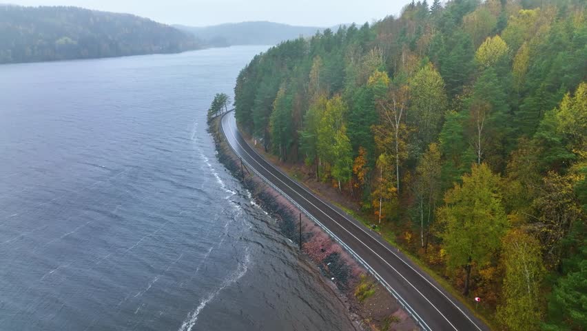 A high angle aerial view of a winding asphalt road running between a vast, dark, and choppy lake or sea and a hillside covered in autumn forest foliage on a cloudy day
