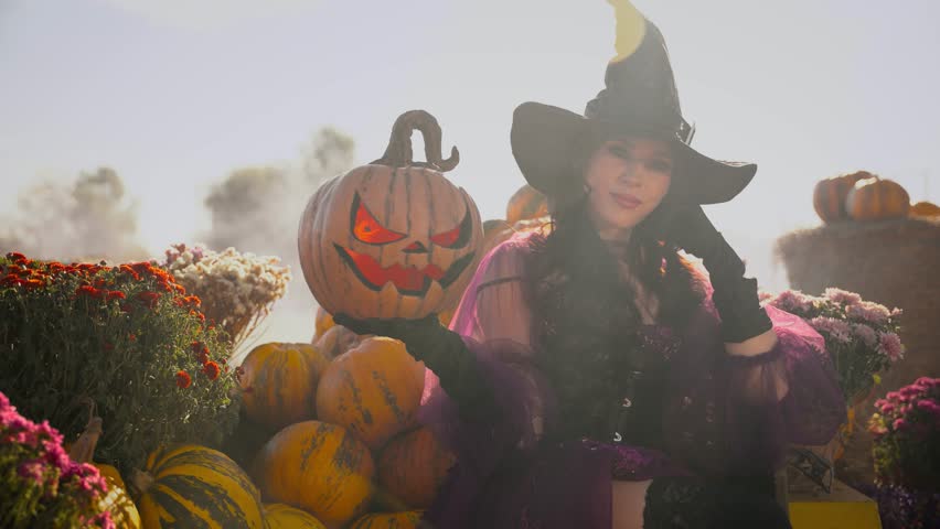 real person happy beauty young woman witch with orange pumpkin carved Jack o Lantern. fantasy smiling face mystery girl in purple dress black hat. Halloween holiday harvest pumpkins Thanksgiving Day.