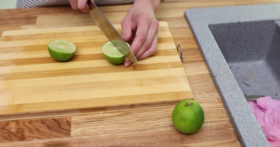 Woman hands cut lime into slices with sharp knife on wooden cutting board. Housewife prepares sour exotic ingredient for cocktails in kitchen
