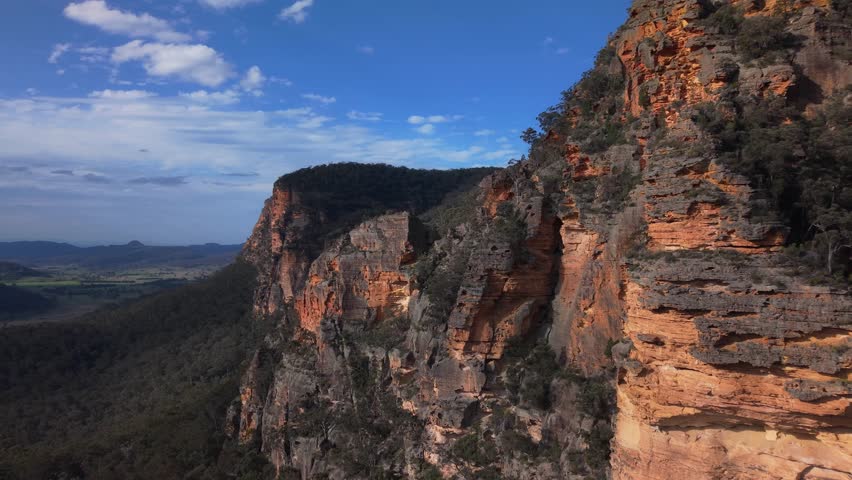 Close drone sweep past sheer sandstone cliffs with deep forest below and open horizon beyond, Blue Mountains NSW Australia
