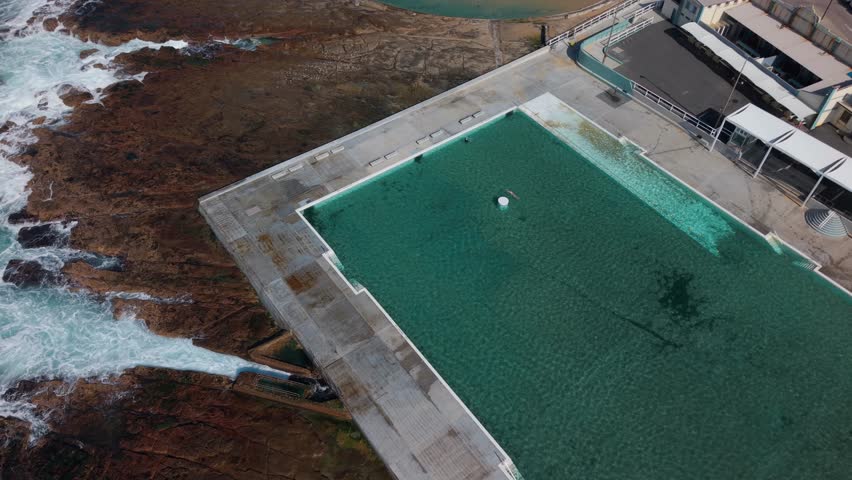 Angled aerial establishing of people enjoying Newcastle Ocean Baths with clean turquoise water and nearby beachline visible