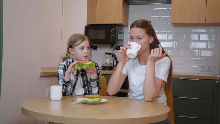 Loving mother and daughter sharing genuine mealtime moment, enjoying homemade sandwiches together at kitchen table, connecting through casual breakfast interaction