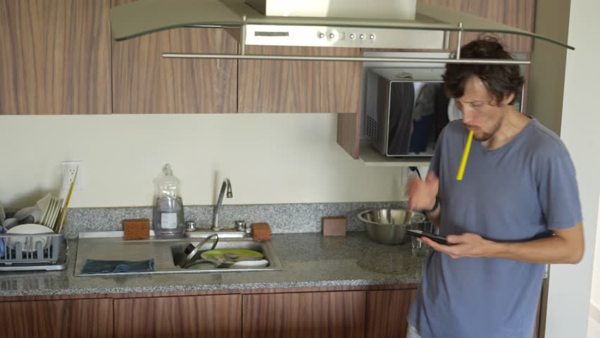 A young man in a kitchen brushes his teeth while engrossed in his phone, showcasing a modern blend of multitasking and phone addiction. A relatable moment reflecting technology's role in daily