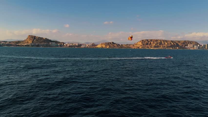 Aerial drone footage of parasailing activity in Alicante, Spain, with Mediterranean coastline, Santa Bárbara Castle, and city skyline in the background