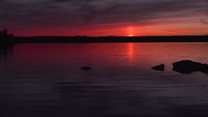 Serene Red Sunset Over a Calm Lake With Silhouetted Rocks, Warm Sky Reflections, and Tranquil Rippling Waters.