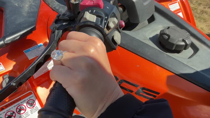 A landscape video tracks steady steering on an orange ATV across rugged ground. The oval diamond engagement ring on a platinum band sparkles through motion and dust.