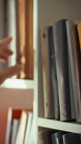 A hand stretches out towards a book on a shelf in a warm, inviting library. The scene captures a moment of exploration and discovery in a quiet space filled with books