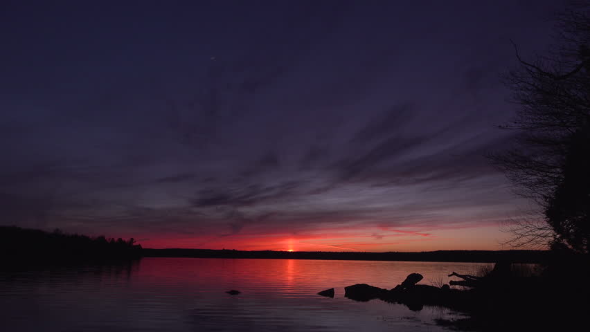Sunset Over A Calm Lake With Silhouetted Rocks, Warm Sky Reflections, Tranquil Water and a Crescent Moon.