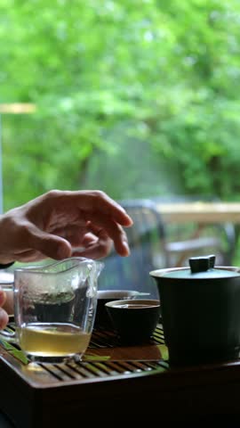 Male hands carefully handling a teacup during a traditional chinese tea ceremony, with a tranquil green garden visible through the window, creating a serene and mindful atmosphere