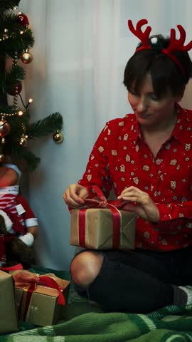 Charming smiling woman in christmas festive clothing is wrapping and decorating Christmas presents while sitting near Christmas tree at home, preparing for New Year 2026. Vertical video