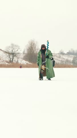 Fisherman with ice drill on shoulder going along ice frozen pond. Man with fishing rod and ice drill is on winter fishing. Vertical video