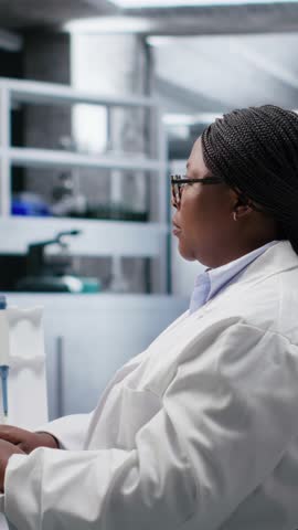 Vertical Video African American woman researcher examining compound in flasks and containers during laboratory testing, studying molecular content with biotechnology equipment. Genetics research