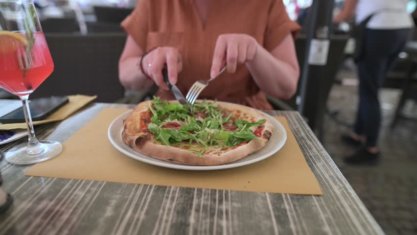 Woman about to eat pizza with arugula and prosciutto, drink next to plate
