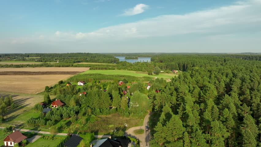A small cluster of rural cottages nestled in a mixed forest patch, overlooking fields and a distant blue lake with an island under a sunny sky - Borzechowo, Poland