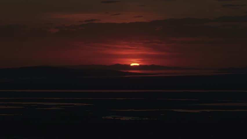 Drone close up shot from Bountiful Canyon in Utah of the sun setting below the Salt Lake Valley with clouds, mountains, and water reflecting light from the Great Salt Lake and a helicopter flying by