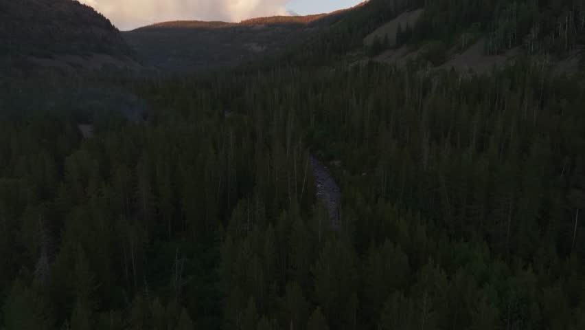 Drone dolly in tilt up wide shot of the Uinta-Wasatch-Cache National Forest in Utah during a summer sunset with the Provo River surrounded by pine trees, and Bald Mountain rising above the valley.
