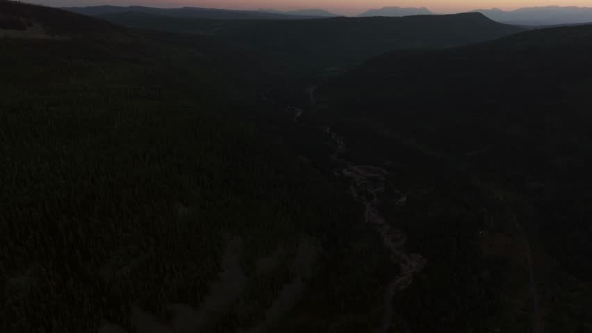 Drone extreme wide tilt up shot of a valley in Uinta-Wasatch-Cache National Forest, Utah, at sunset with the Provo River surrounded by pine trees, Highway 150 to the right, mountains and a summer sky.