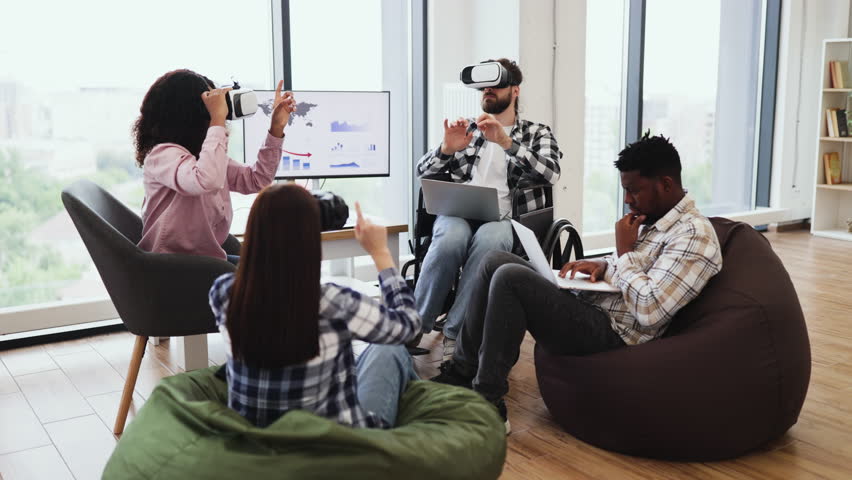 Young adults engage with virtual reality devices and laptops in collaborative office. Diverse teams demonstrating technology interaction seated on floor and chairs creating innovative atmosphere.