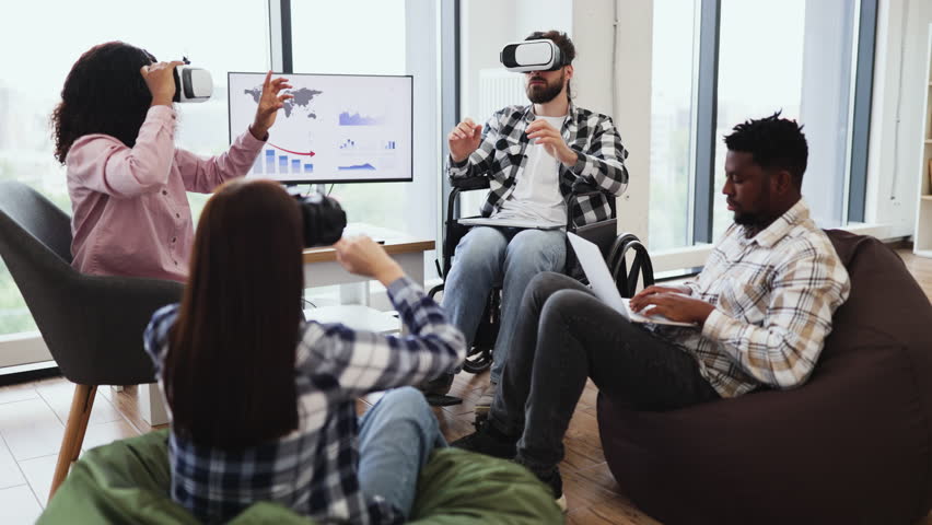 Diverse group of young adults including male wheelchair user enjoying virtual reality. Another male working on laptop sitting on bag chair in modern office setting - Powered by Shutterstock - Get 15% off with code: PIKWIZARD15