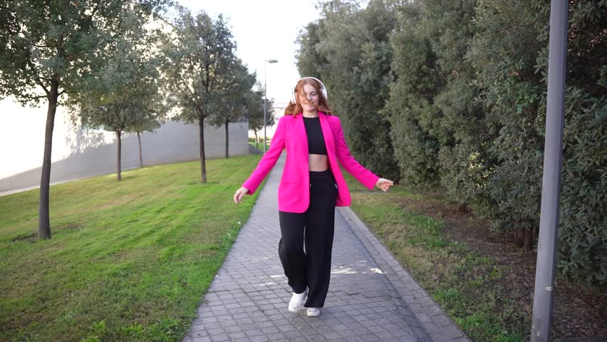 A fashionable woman in a chic pink blazer and stylish black pants strolls along a scenic pathway lined with trees, proudly showcasing her vibrant style in this serene outdoor setting