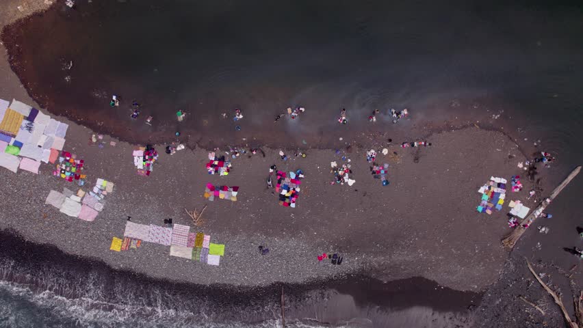 Praia Piscina, São Tomé and Príncipe - 2nd September 2025: A large group of women washing clothes in a river opposite a scenic beach on São Tomé island.