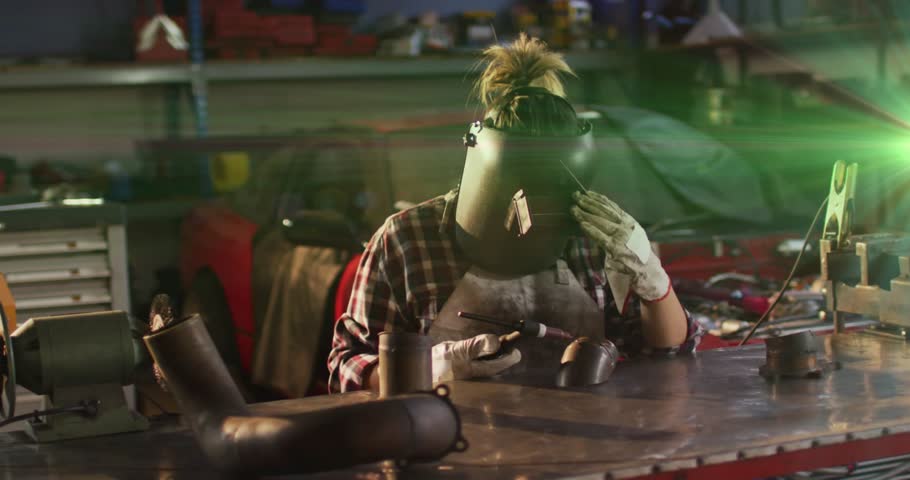 Female welder picking up helmet and welding steel exhaust pipe with sparks in automotive workshop. Industrial, craftsmanship, metalwork, manufacturing, precision, safety, technology