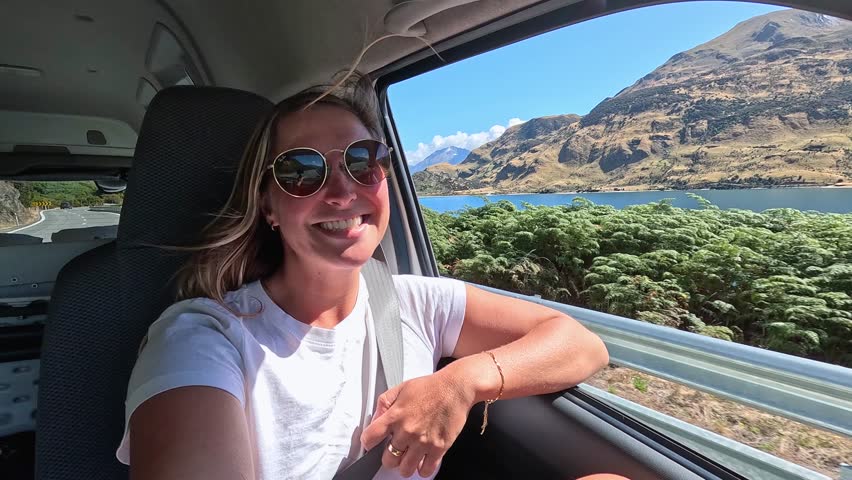 A girl in a camper van passenger seat gazes at Lake Wanaka, South Island, New Zealand, as they drive past. The calm lake, mountains, and greenery make for a scenic journey.